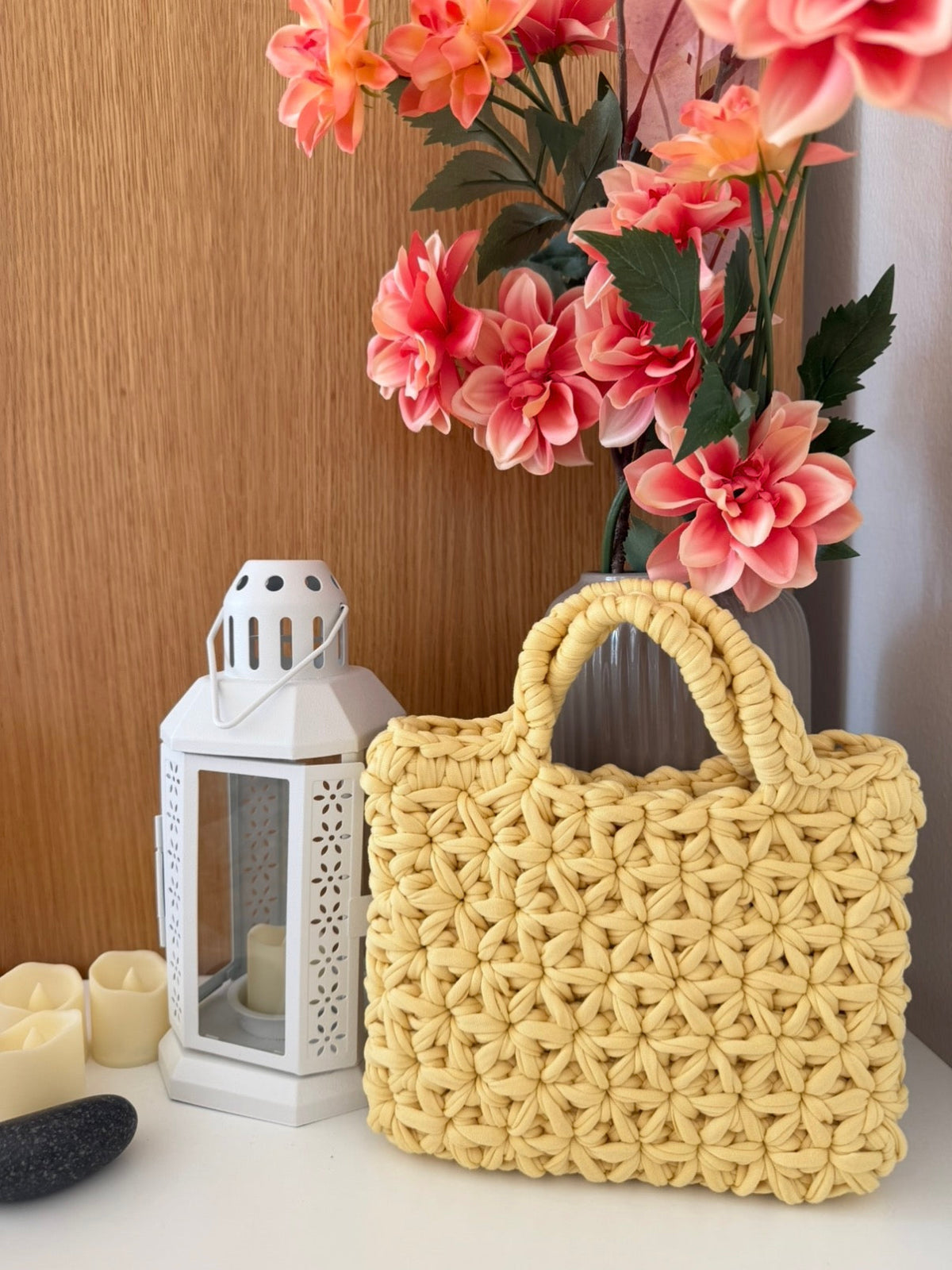 A yellow woven straw handbag with double top handles sits on a white surface next to a white lantern, with pink flowers in a vase and a wooden wall in the background.