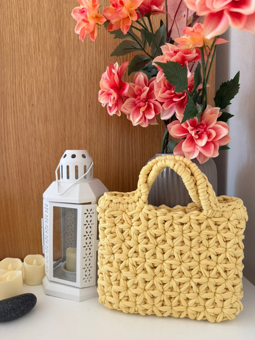 A yellow woven straw handbag with double top handles sits on a white surface next to a white lantern, with pink flowers in a vase and a wooden wall in the background.