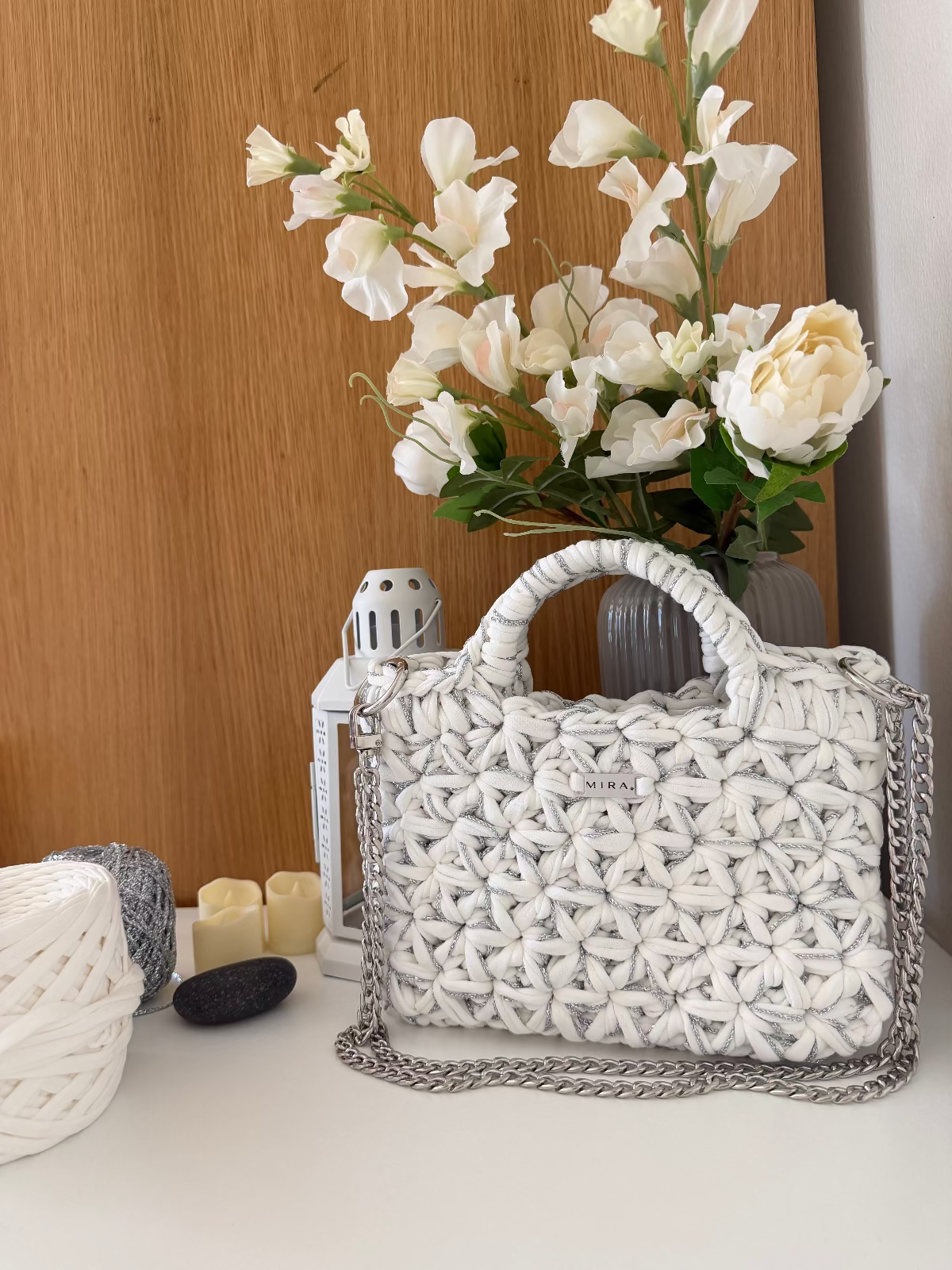 A white woven textured handbag with a silver chain strap, styled on a tabletop next to a vase of white flowers, candles, and yarn balls.