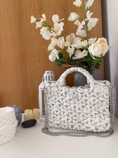 A white woven textured handbag with a silver chain strap, styled on a tabletop next to a vase of white flowers, candles, and yarn balls.