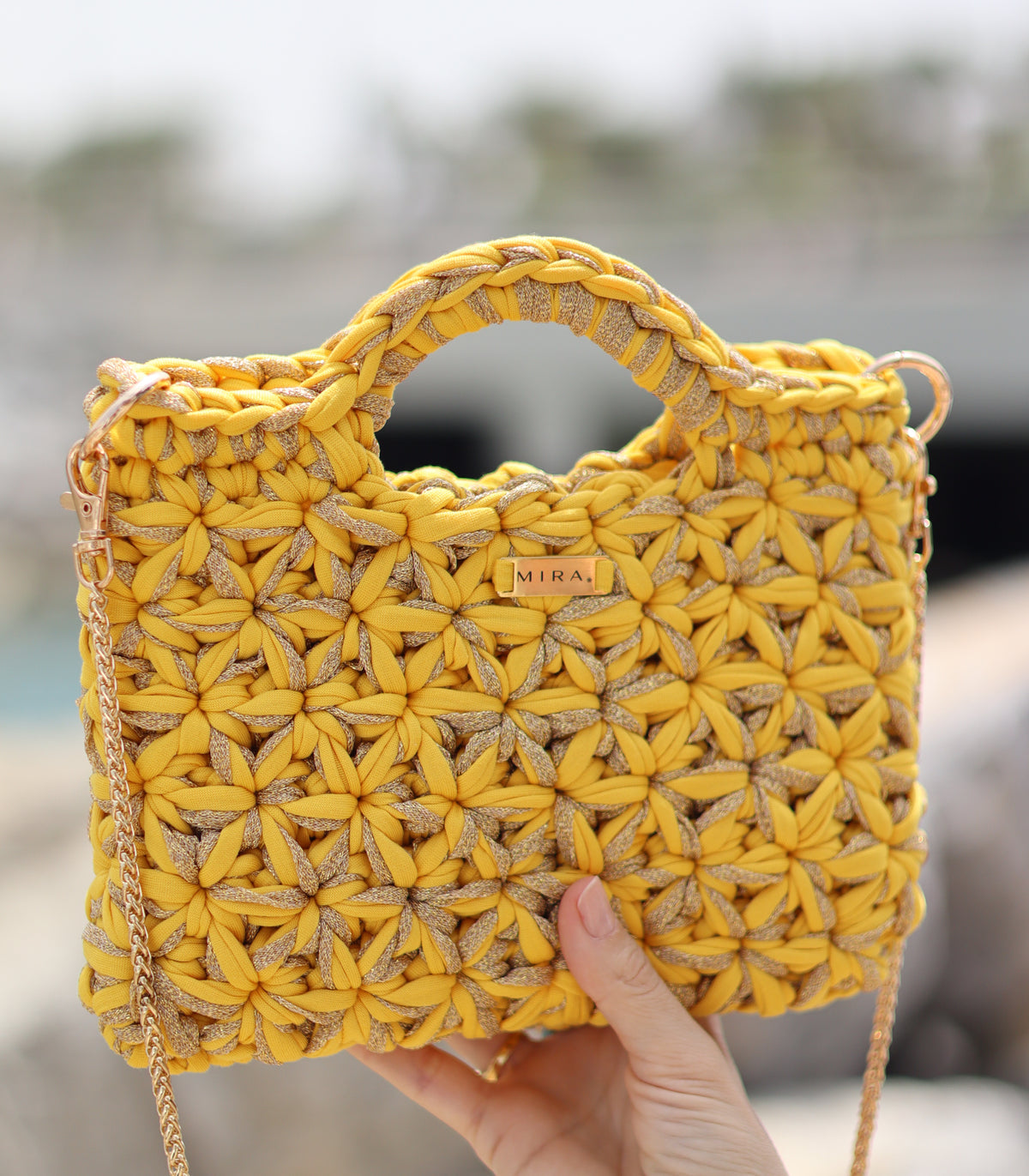 A yellow and beige chunky crocheted handbag with a top handle and gold chain strap, held by a hand against a blurred outdoor background.